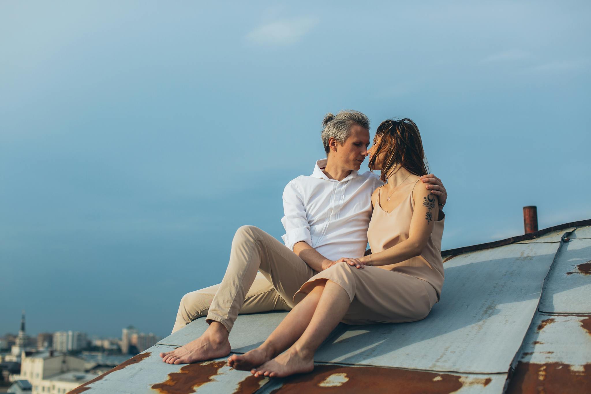 A couple embraces on a rooftop, enjoying a romantic moment at sunset with a cityscape background.