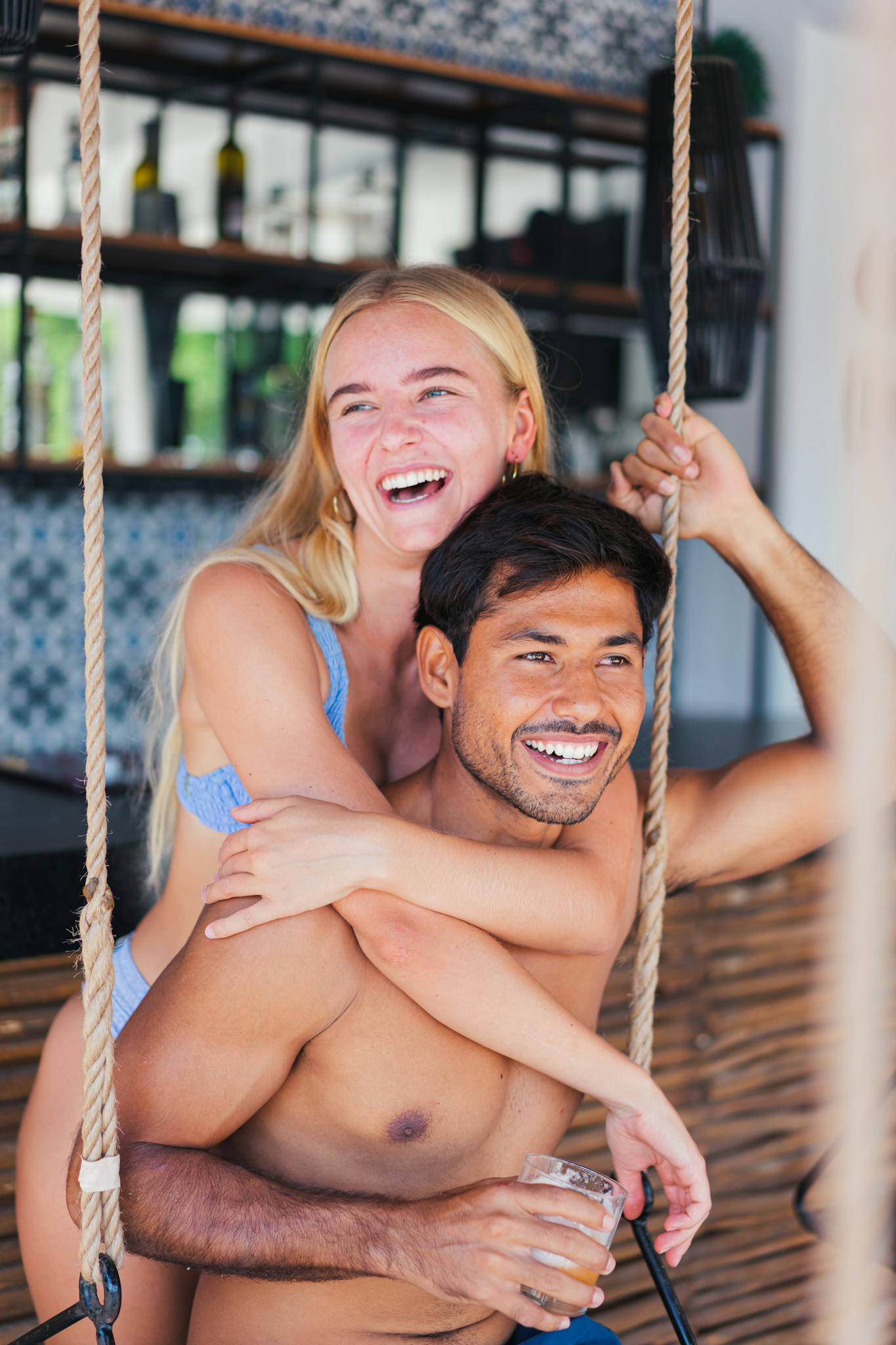 Young couple laughing and embracing on a swing at an outdoor beach bar, enjoying a sunny day.